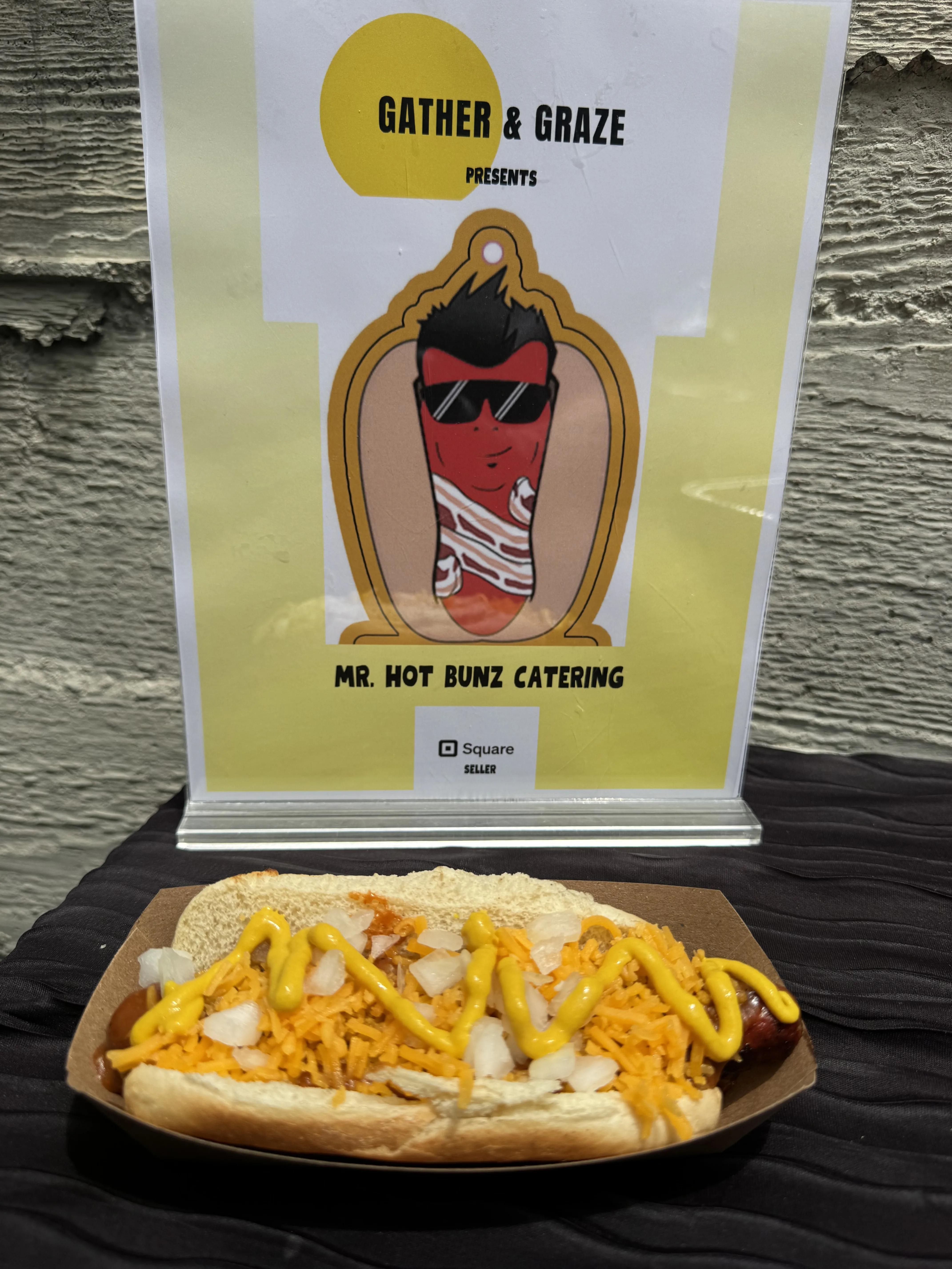 Loaded bacon-wrapped hot dog with cheese, onions, and mustard at a Gather & Graze event